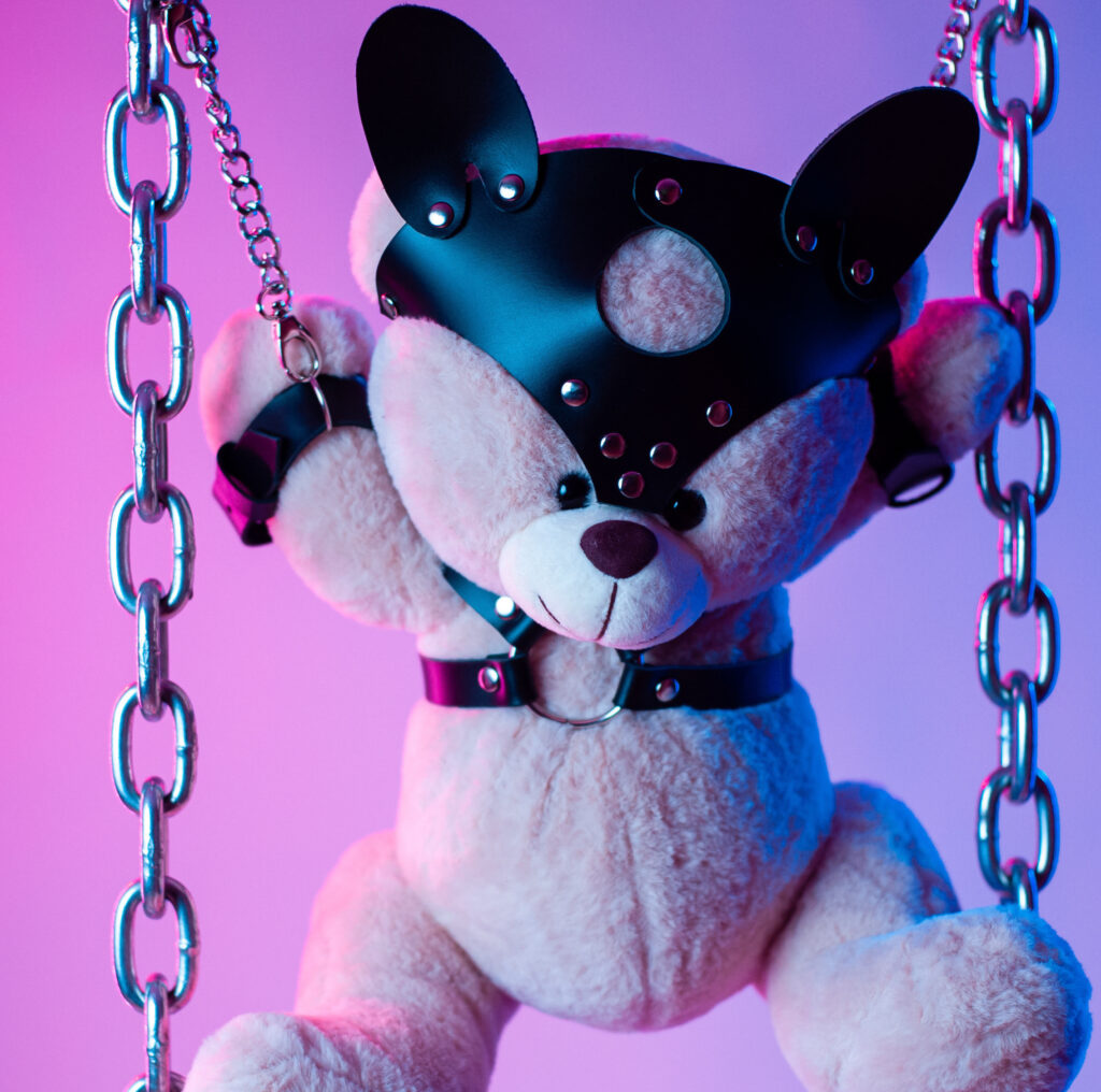 toy bear dressed in leather belts harness accessory for BDSM games on a dark background in neon light By rotozey